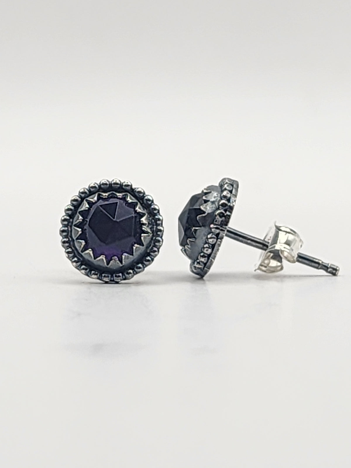 A pair of round stud earrings featuring a faceted dark purple center stone surrounded by a textured silver bezel and beaded border, with one earring shown from the front and the other from the side displaying the post and butterfly backing.
