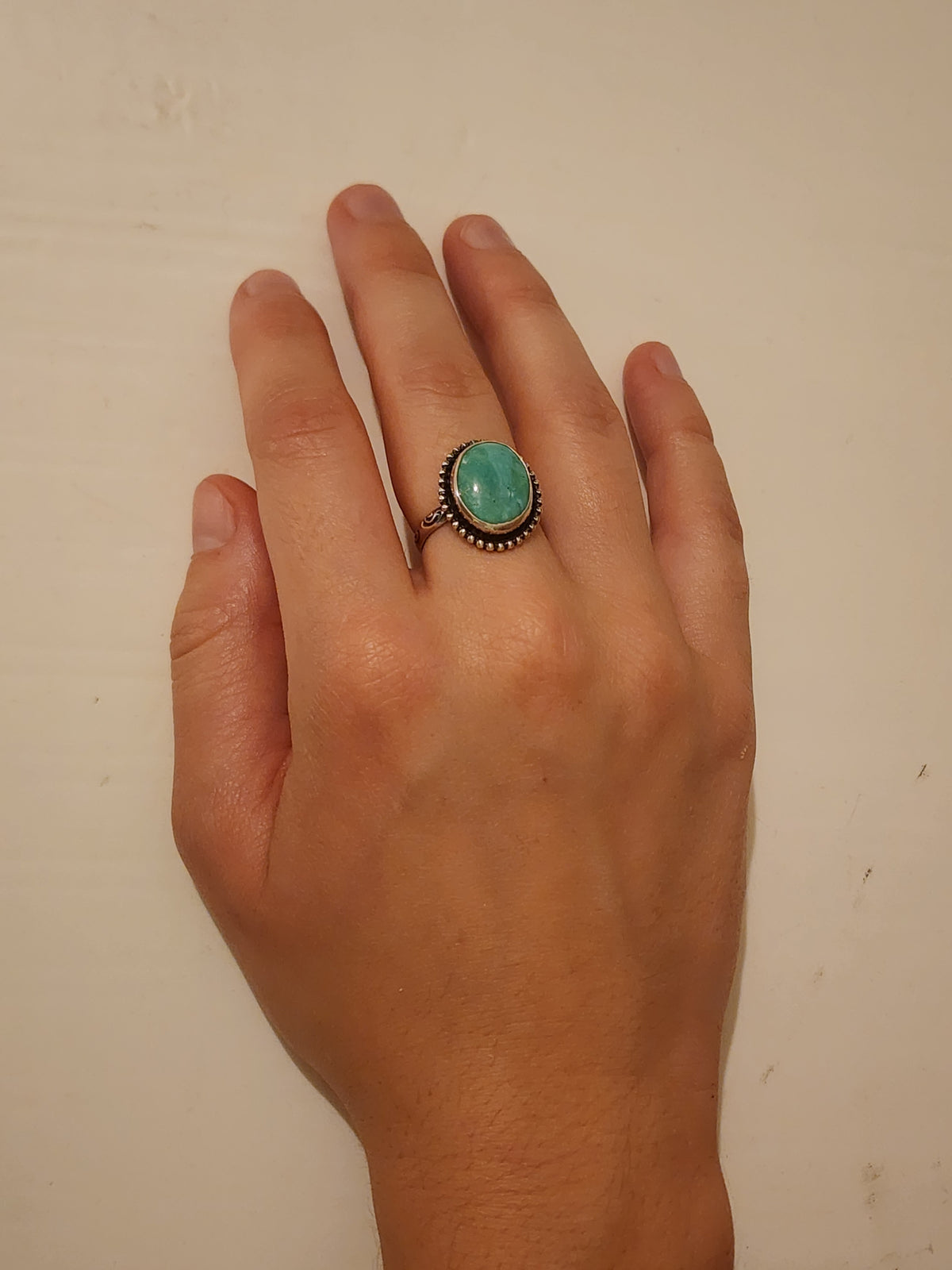 Sterling silver ring with oval turquoise stone and beaded border displayed on the middle finger of a hand on an off white background.