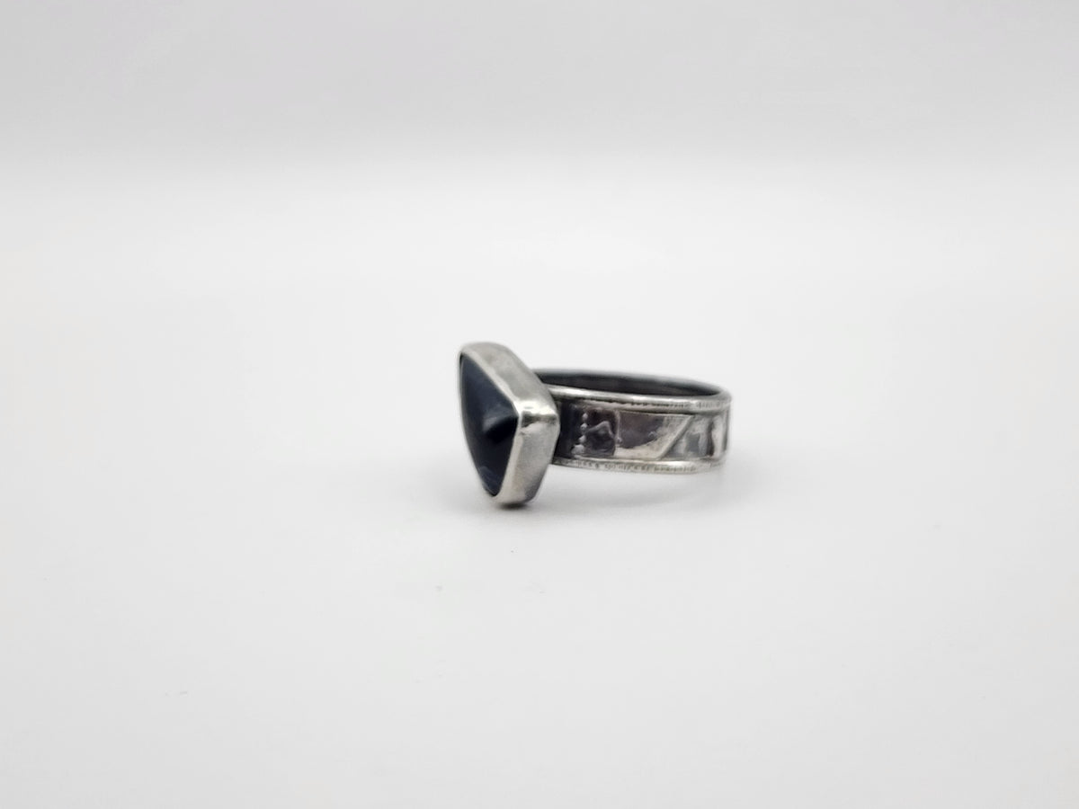 A sterling silver ring with a irregular triangular dark stone offset on a textured band, photographed on a white background.