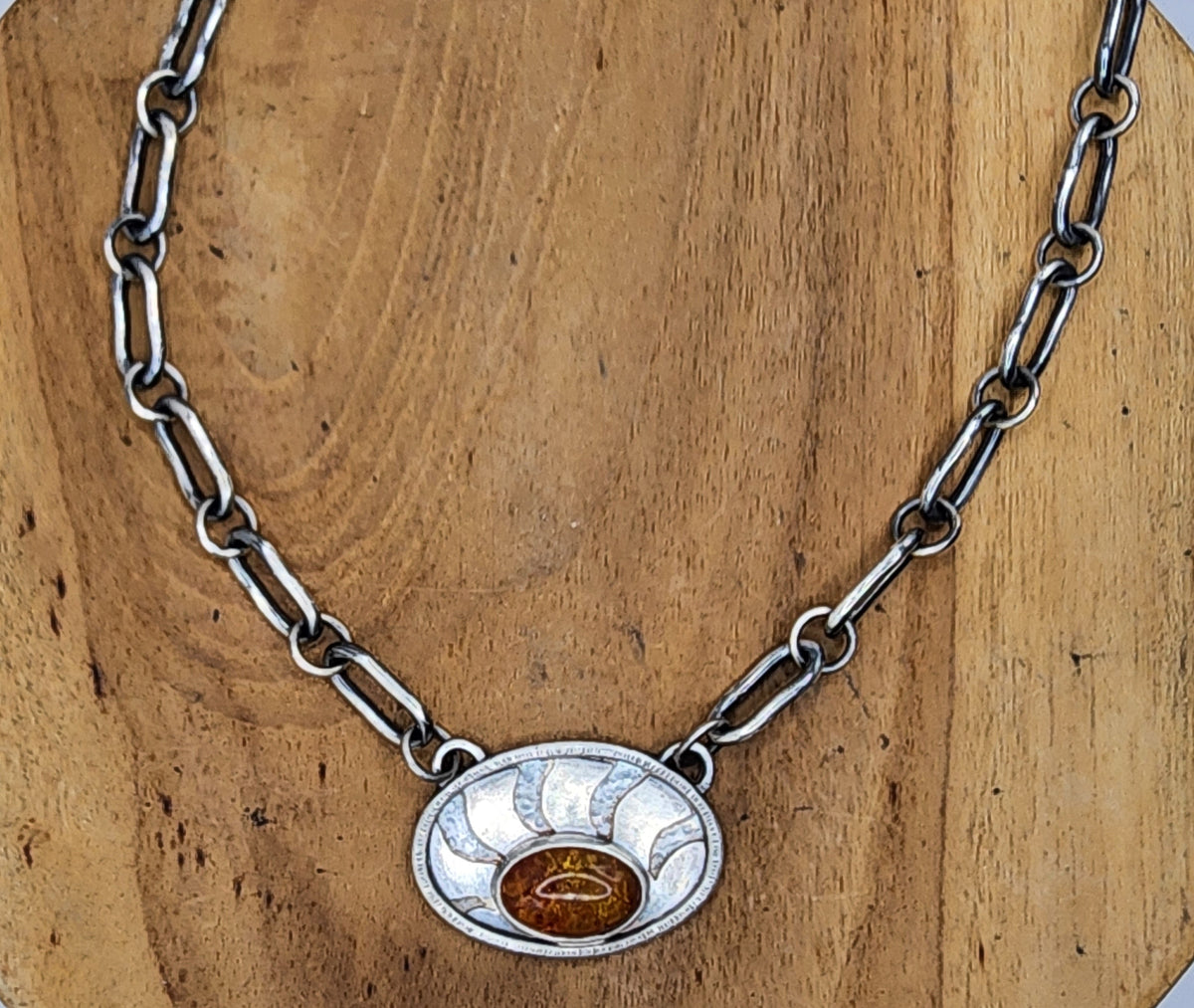 A sterling silver link chain necklace with an oval pendant featuring sunrays and an oval baltic amber in the lower center, displayed on a round wooden stand.
