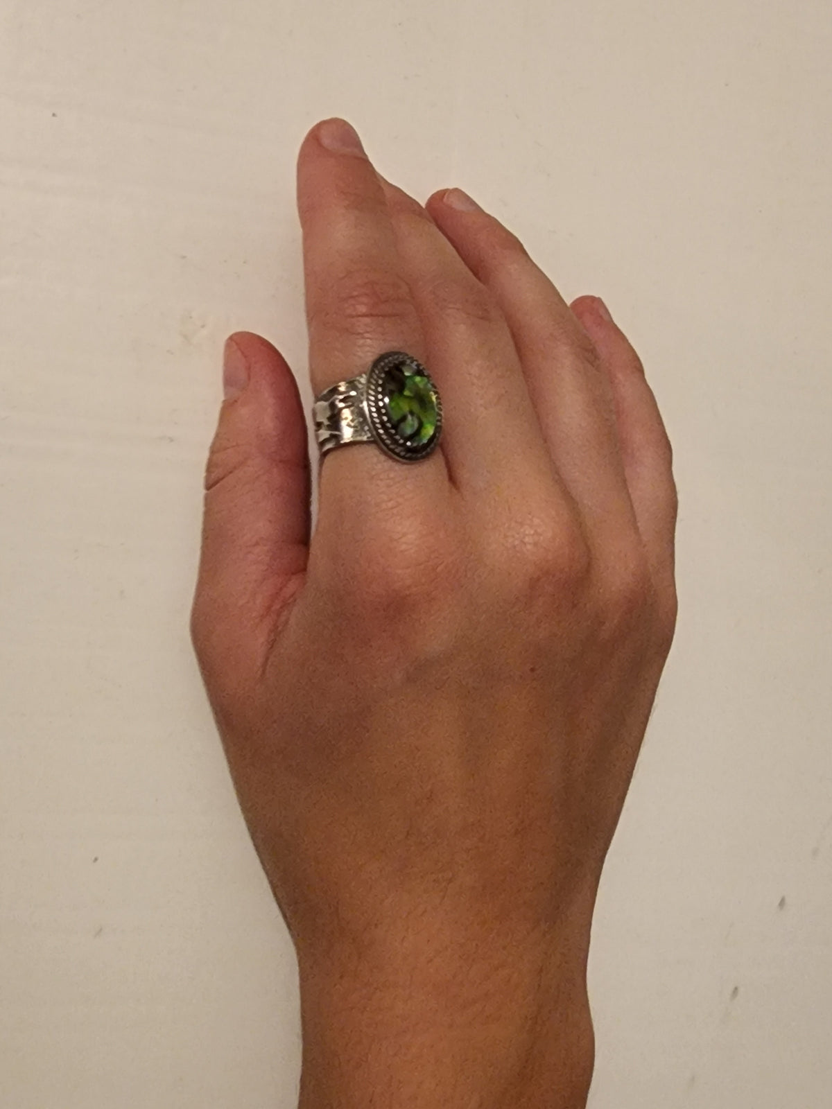 A sterling silver ring with gator on wide band and oval abalone with twisted band wire on a person's pointer finger on an off white background.