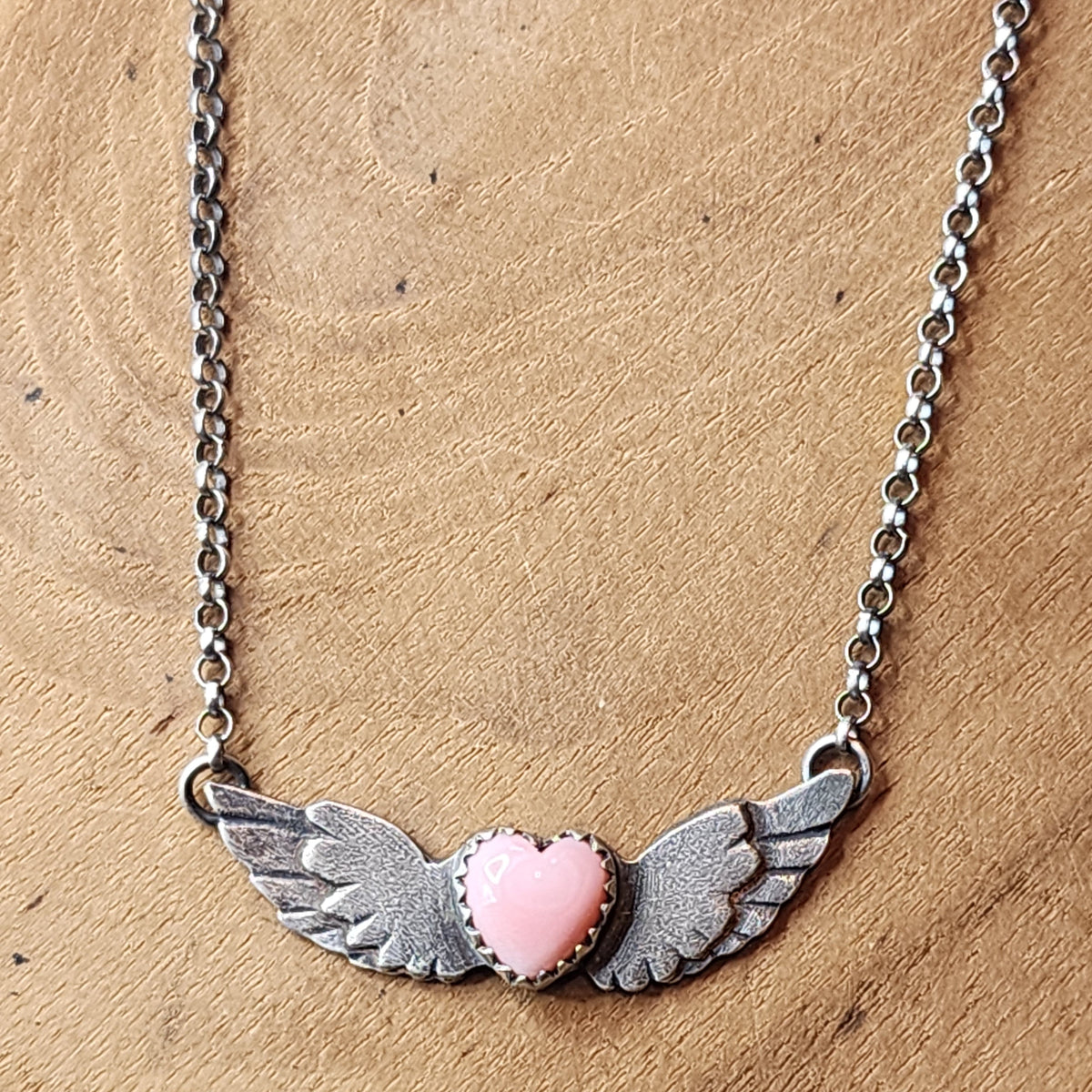 A sterling silver winged heart pendant necklace with a pink heart in the center, displayed on a textured brown wooden surface.