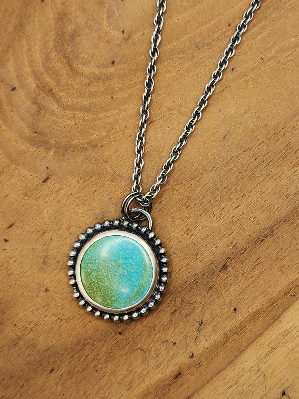 Sterling silver pendant with round turquoise cabochon and beaded wire border on silver chain, sitting on wooden background.