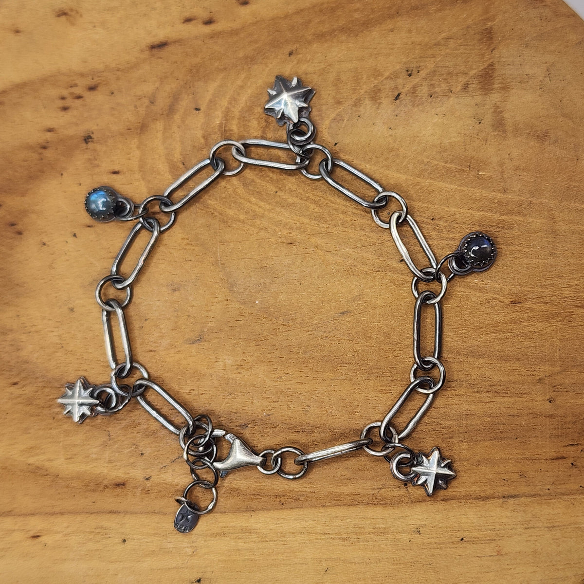 A sterling silver paperclip chain bracelet with three star and two labradorite charms, displayed on a round wooden surface.
