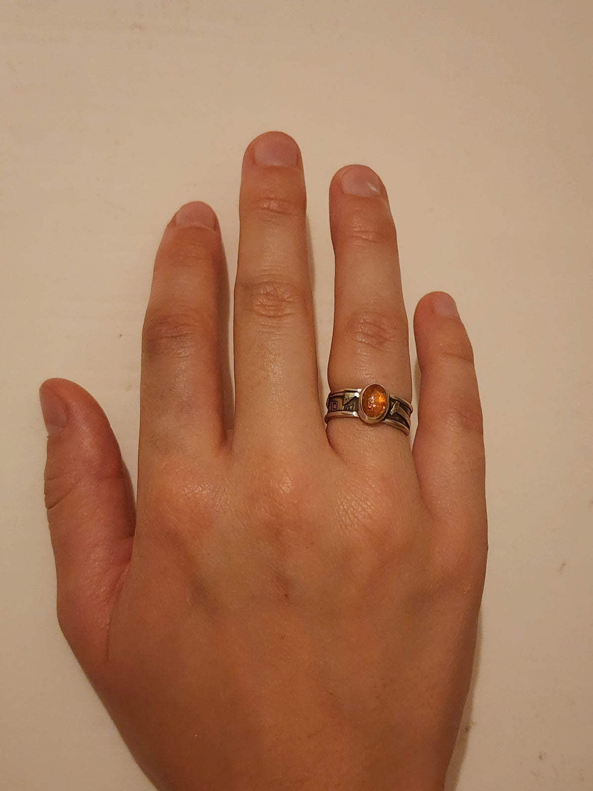 A sterling silver ring with heavily textured band and round orange stone on the ring finger of a person's hand on an off white background.