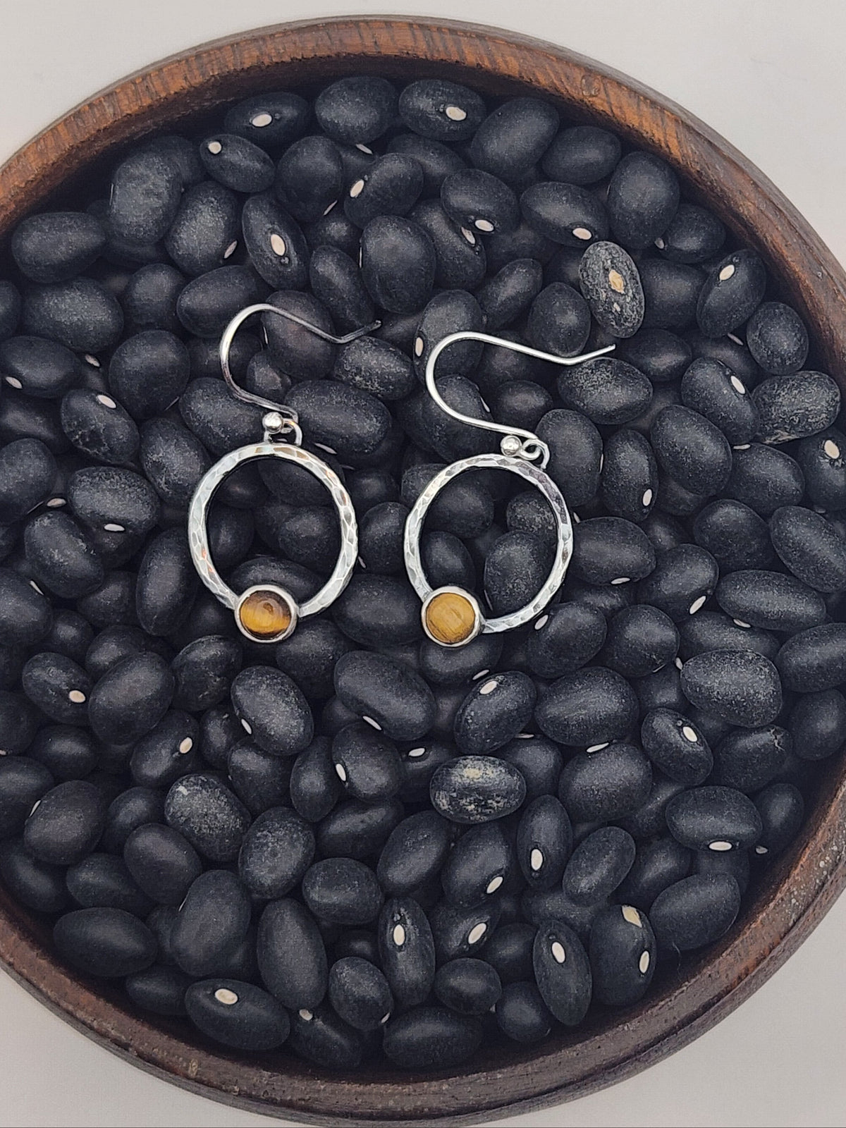 A pair of sterling silver hoop dangle earrings with small round tigers eye stones, displayed on a bed of black beans inside a wooden bowl against a light gray background.