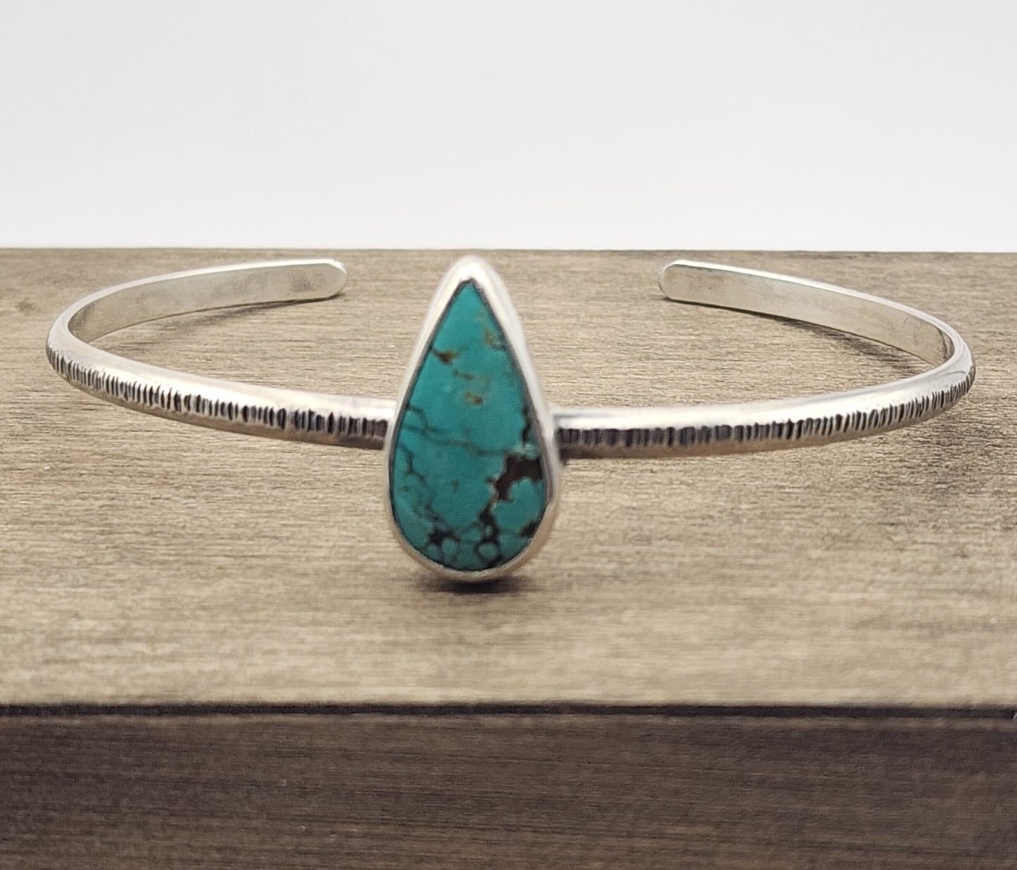 A sterling silver textured cuff bracelet featuring a teardrop-shaped turquoise stone in the center, displayed on a wooden surface.