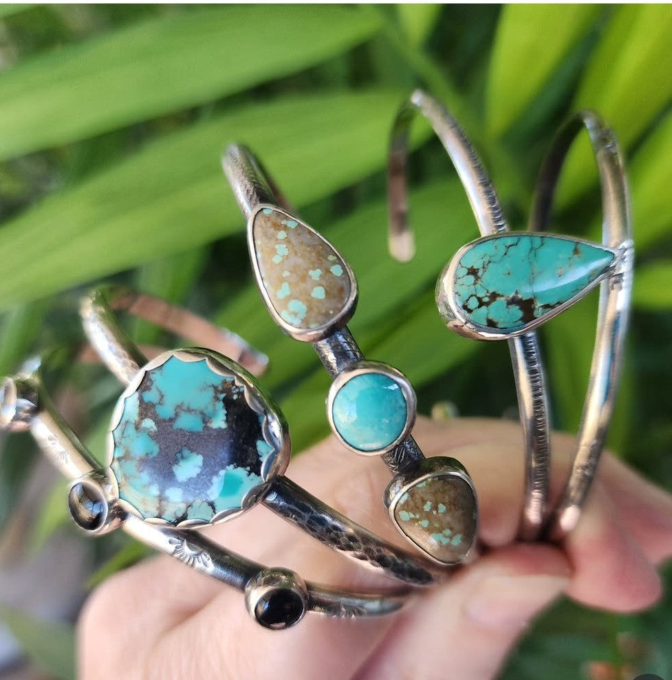 Hand holding 5 cuff bracelets with gemstones and plant in background