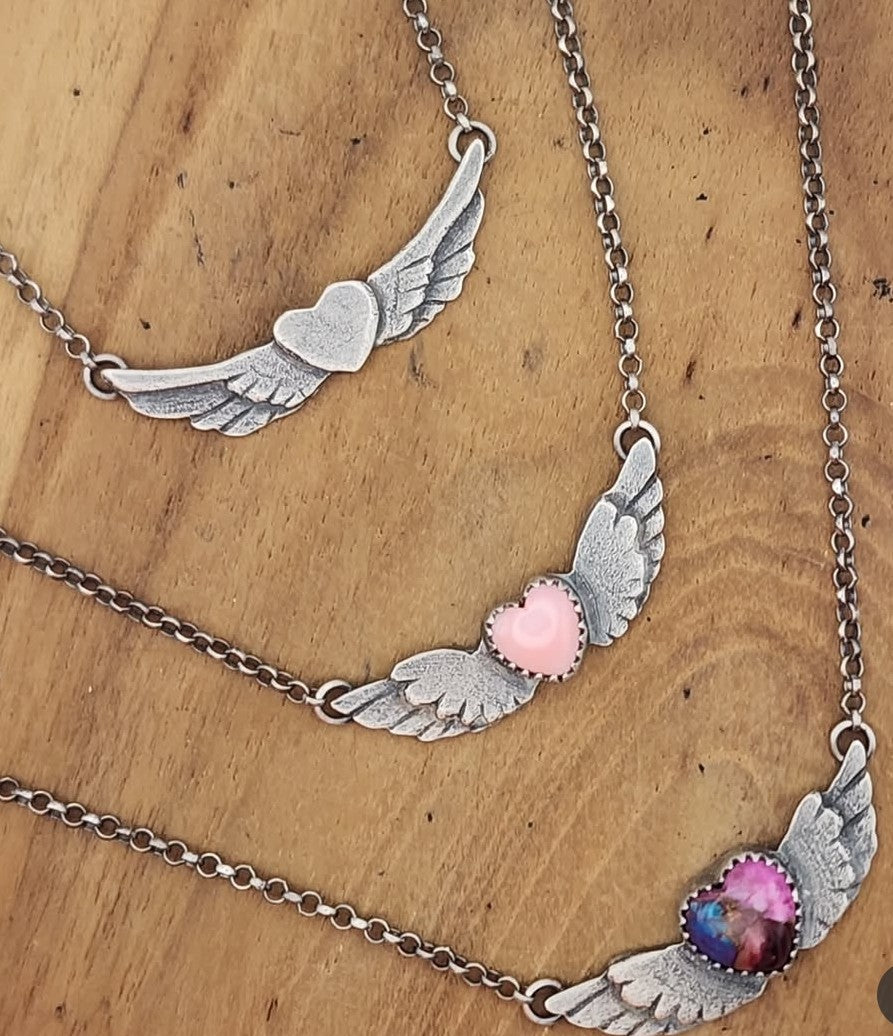 Three necklaces with hearts and wings, one with no stone, one with pink heart and one with pink and purple heart on a wooden background