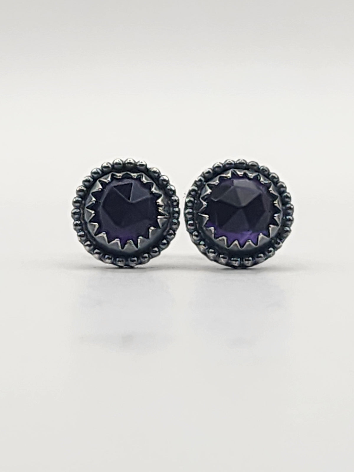 A pair of round stud earrings featuring rose cut dark purple gemstones in a textured, serrated bezel setting with a beaded border, shown on a light gray reflective surface.