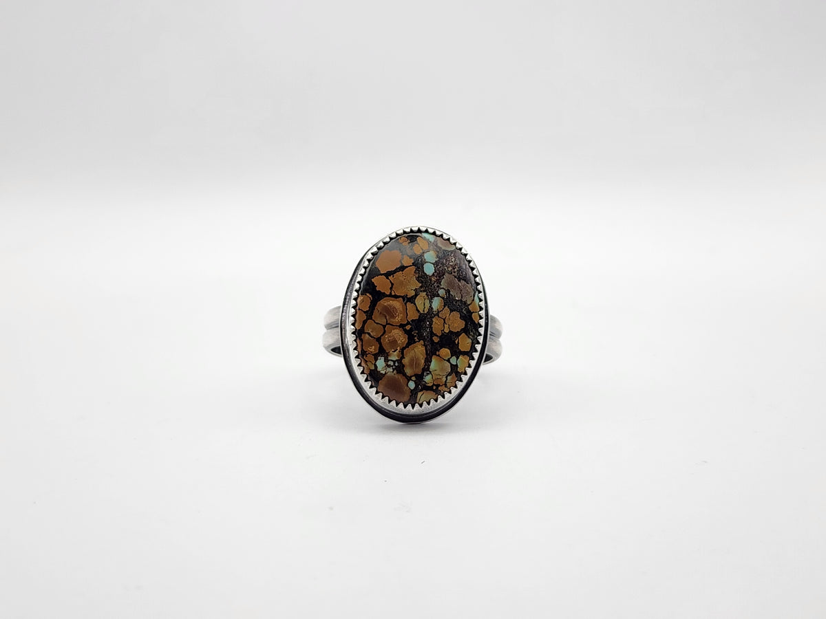A sterling silver ring featuring a large oval cabochon stone with a brown, black, and orange matrix pattern and small turquoise flecks, set against a plain light gray background.
