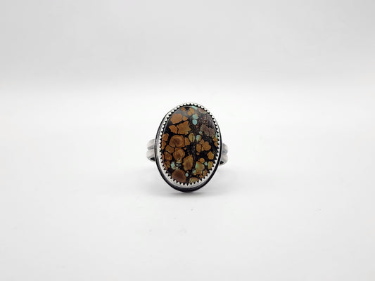 A sterling silver ring featuring a large oval cabochon stone with a brown, black, and orange matrix pattern and small turquoise flecks, set against a plain light gray background.