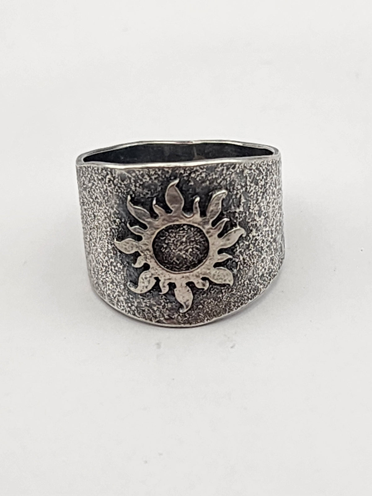A wide textured sterling silver band with a raised sun image on the center on a white background
