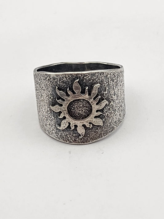 A wide textured sterling silver band with a raised sun image on the center on a white background