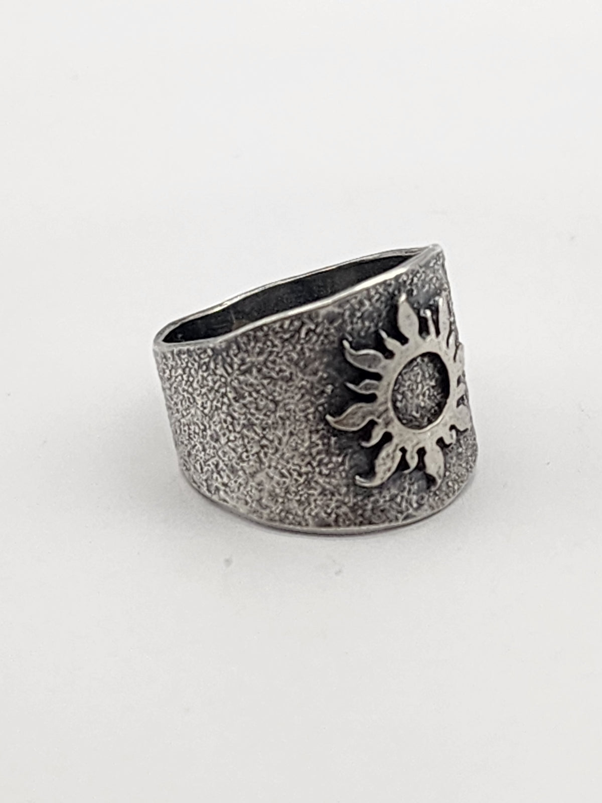 A wide sterling silver textured band, side view of ring with raised sunburst in center of ring. Ring is on a white background. 
