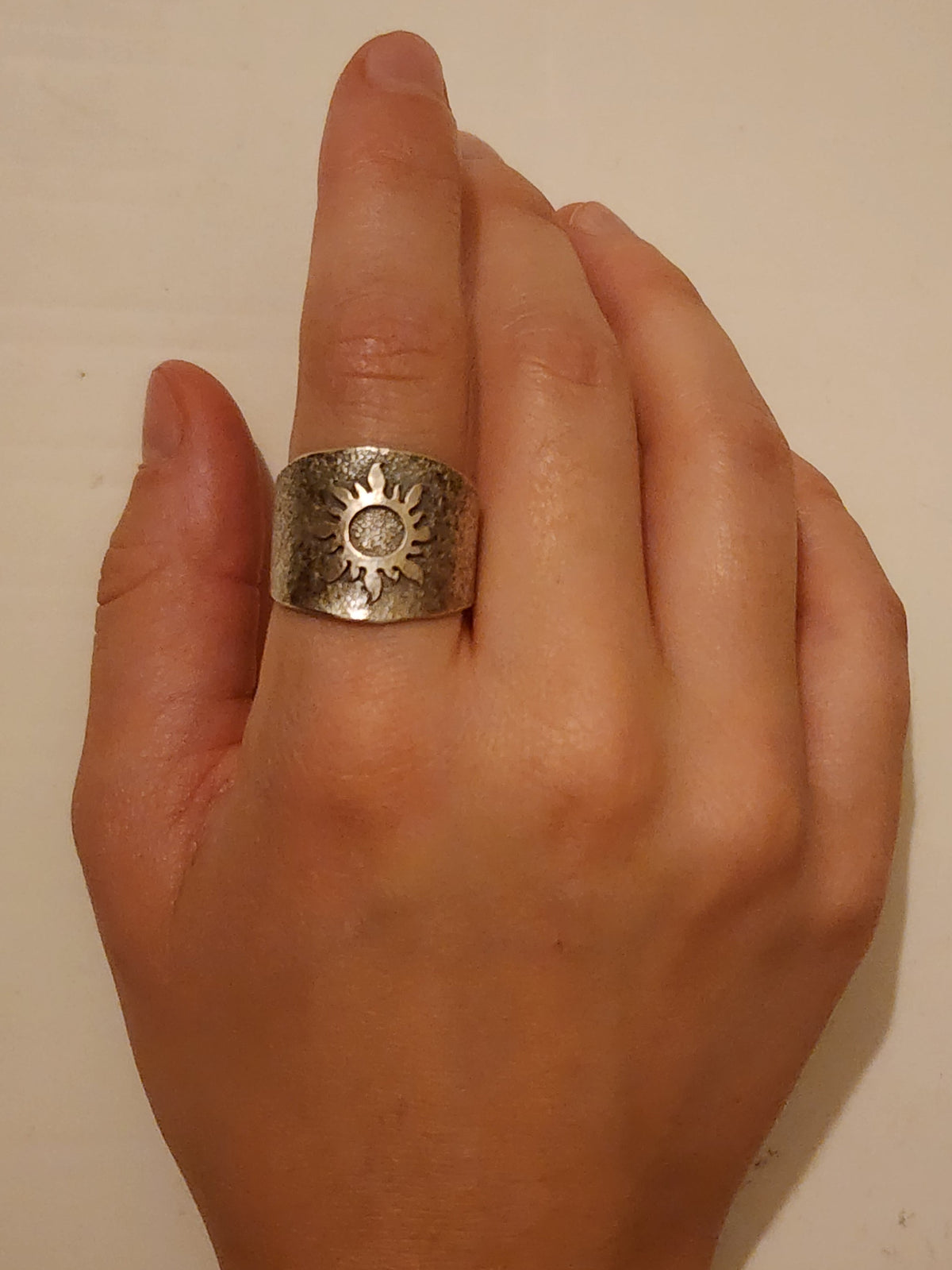 A wide textured sterling silver ring with a sunburst in the middle on a person's pointer finger on an off white background. 
