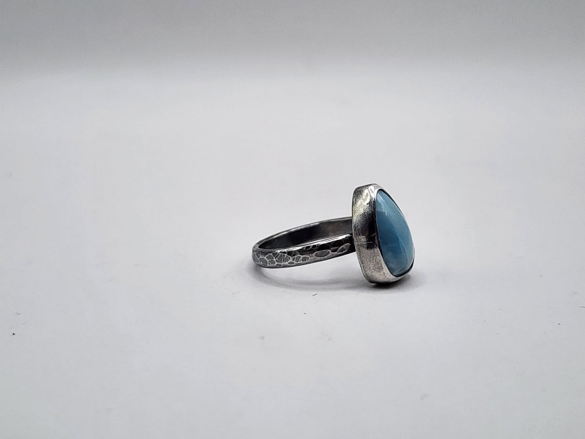 A sterling silver ring featuring a teardrop-shaped light blue gemstone on a hammer textured ring band, photographed on a neutral light gray background.
