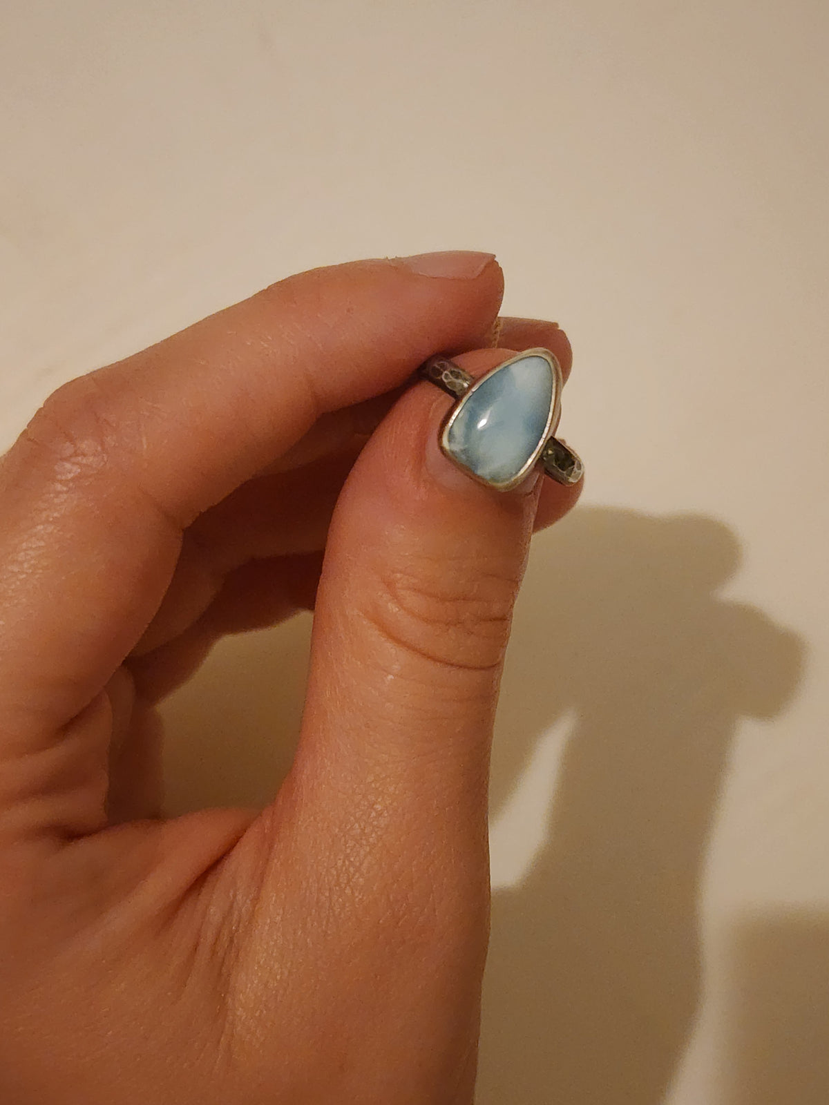 A hand holding a silver ring with a light blue teardrop-shaped gemstone against a plain beige background.
