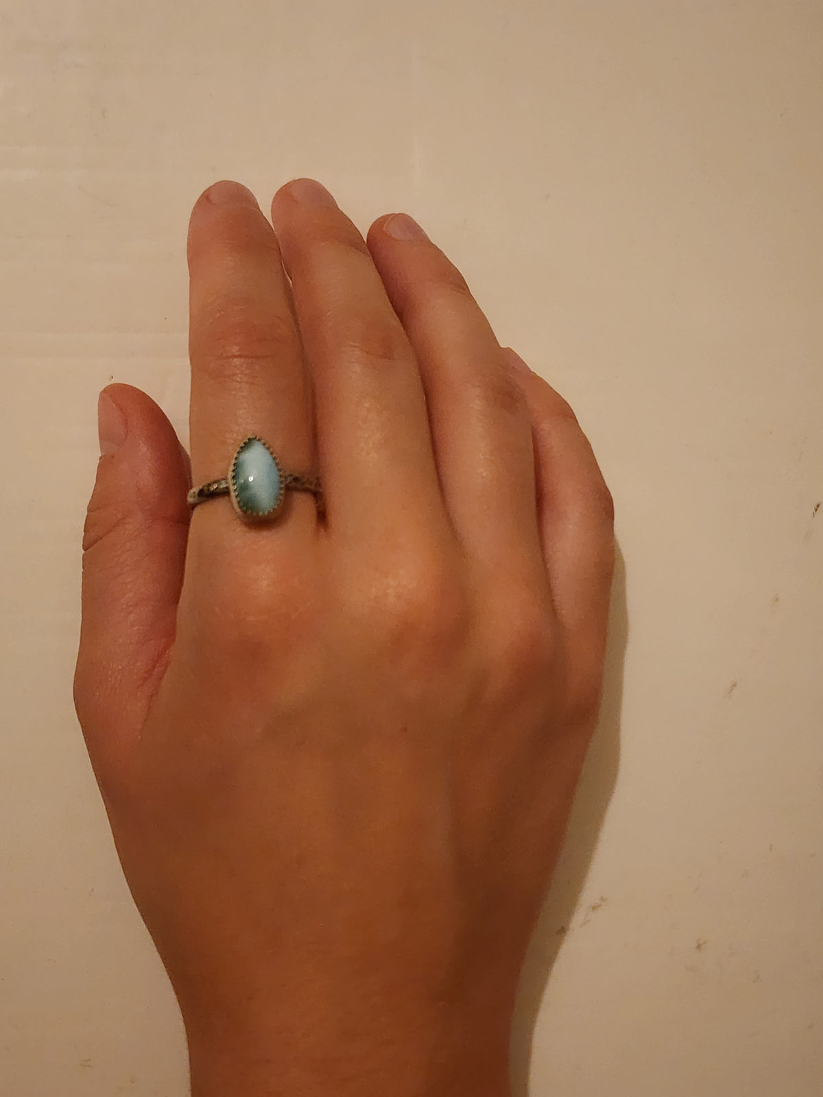 A sterling silver ring with a blue teardrop gemstone on the pointer finger of a hand photographed on an off white background.