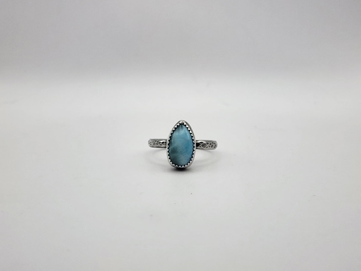 A sterling silver ring with teardrop blue stone and serrated bezel set on a hammer textured ring and oxidized, photographed on a white/light grey background. 