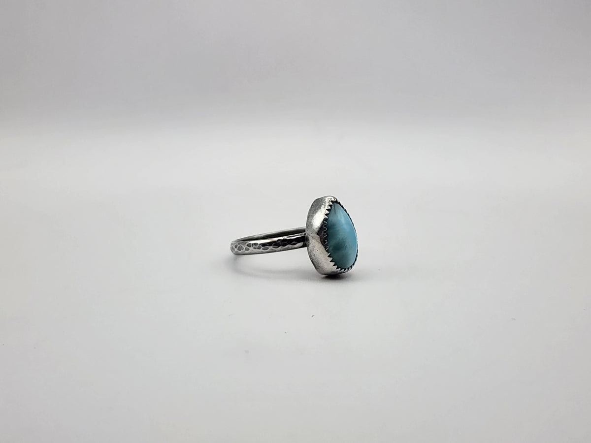 A side view of a sterling silver ring with teardrop blue stone and serrated bezel set on a hammer textured ring and oxidized, photographed on a white/light grey background. 