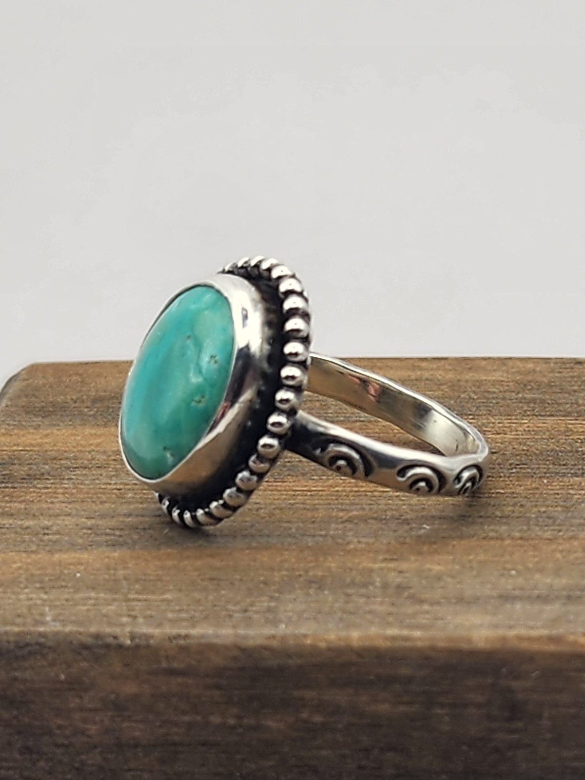 Sterling silver ring with oval turquoise and beaded border with stamped band. Side view of ring sitting on wooden block and white background. 