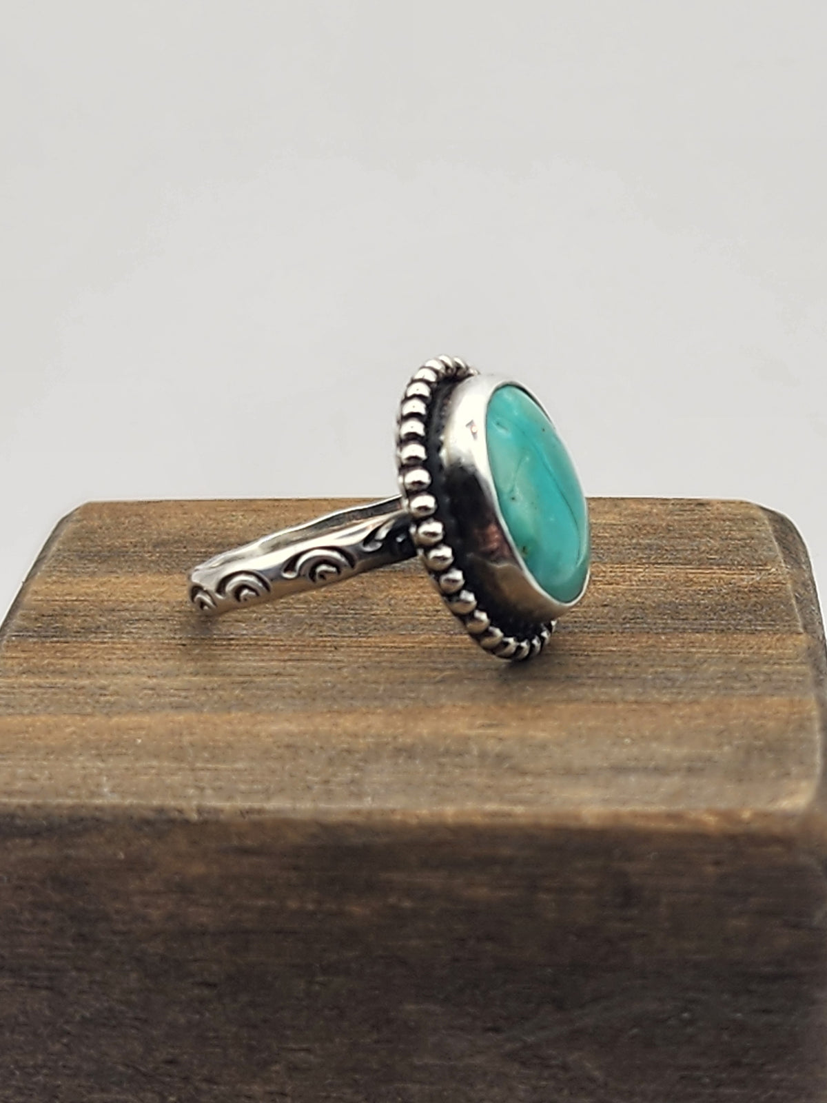 Side view of sterling silver ring with oval turquoise stone and beaded border with stamped ring band. Displayed on a wooden block with white background. 