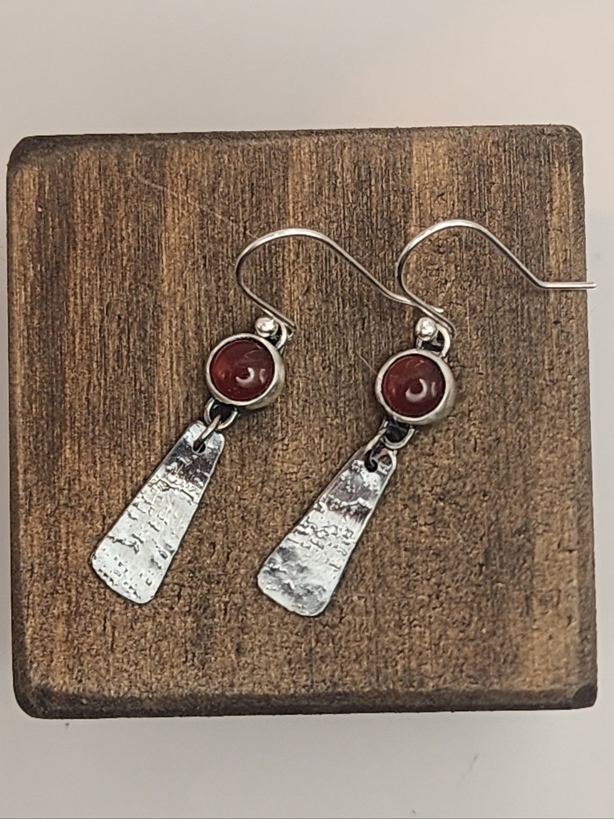 A pair of handmade dangle earrings with textured silver and round carnelian gemstone accents, displayed on a square wooden block against a light neutral background.