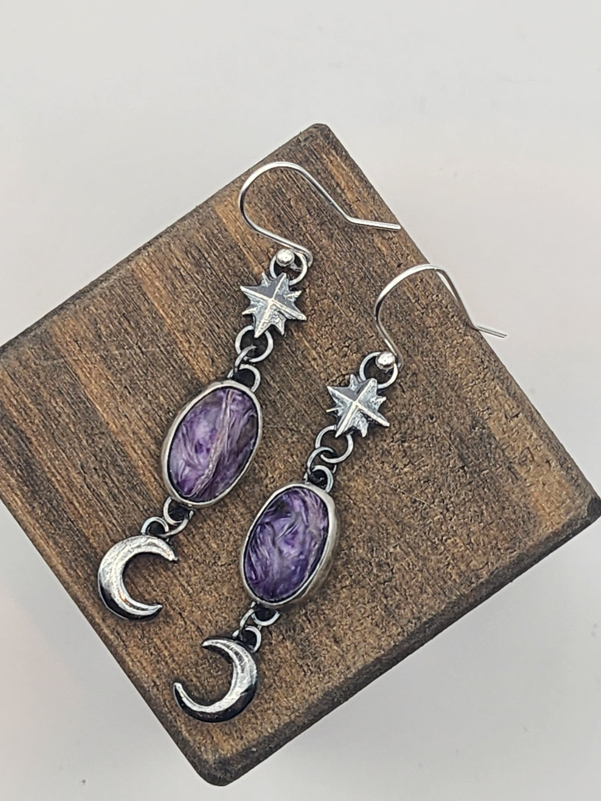 A pair of sterling silver dangle earrings with oval purple gemstones, star charms, and crescent moon charms, displayed on a small wooden block against a light gray background.