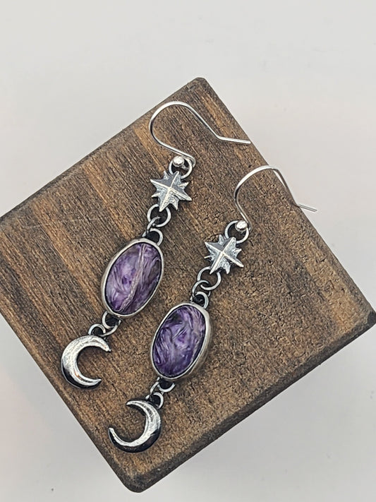 A pair of sterling silver dangle earrings with oval purple gemstones, star charms, and crescent moon charms, displayed on a small wooden block against a light gray background.