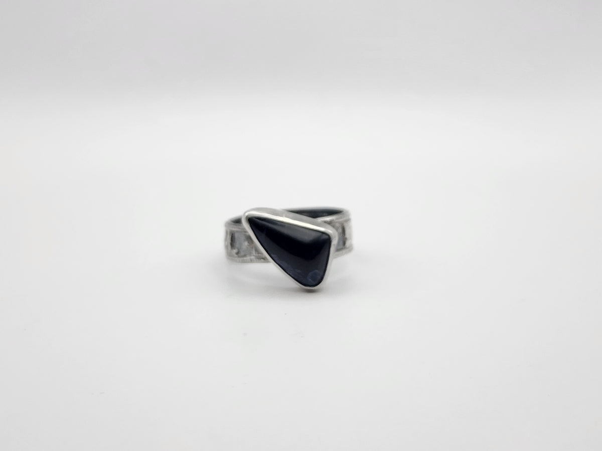 A sterling silver ring with a irregular triangular dark stone offset on a textured band, photographed on a white background. 