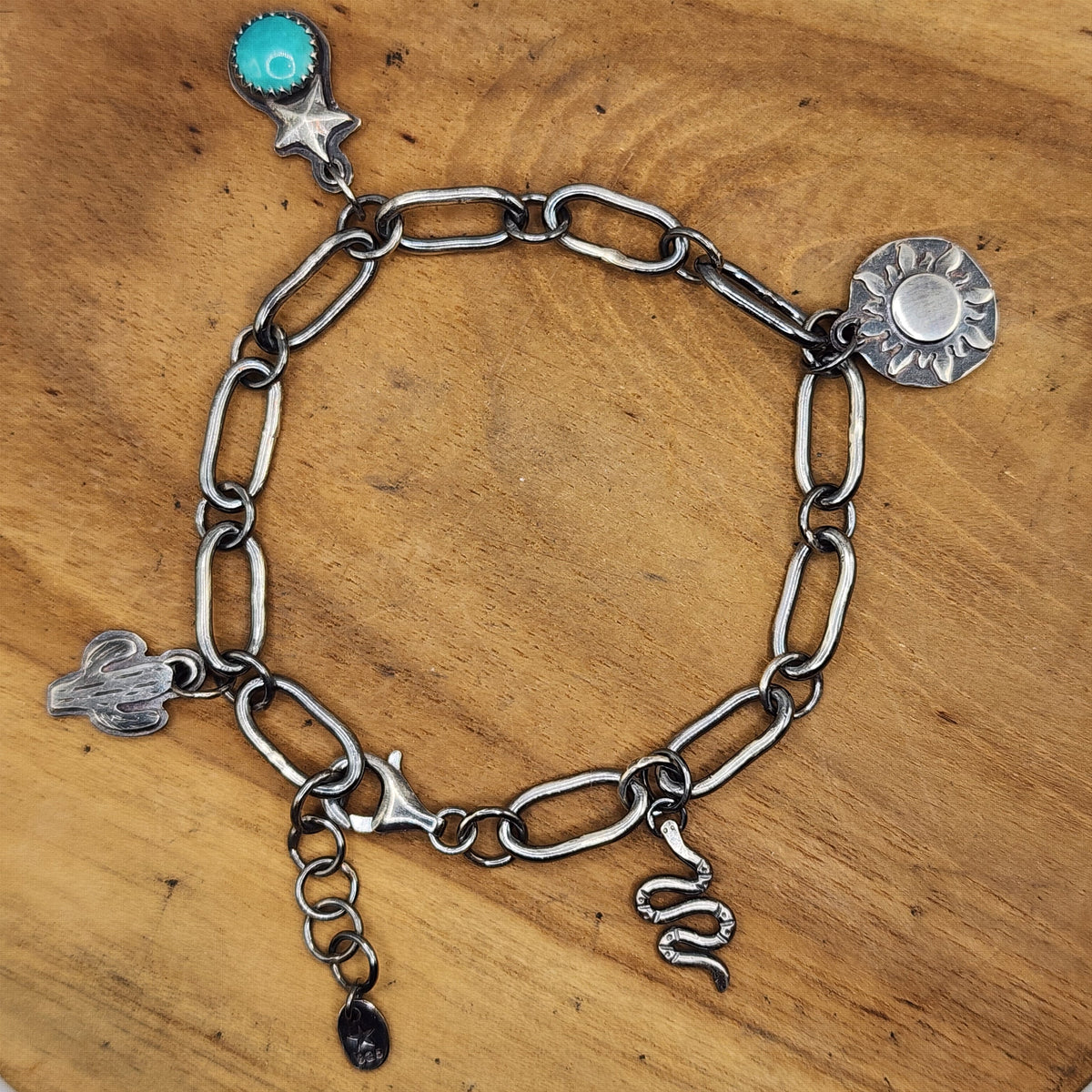 A sterling silver paperclip chain bracelet with various charms, including a turquoise stone star, a sun, a cactus, a snake, displayed on a round wooden board.