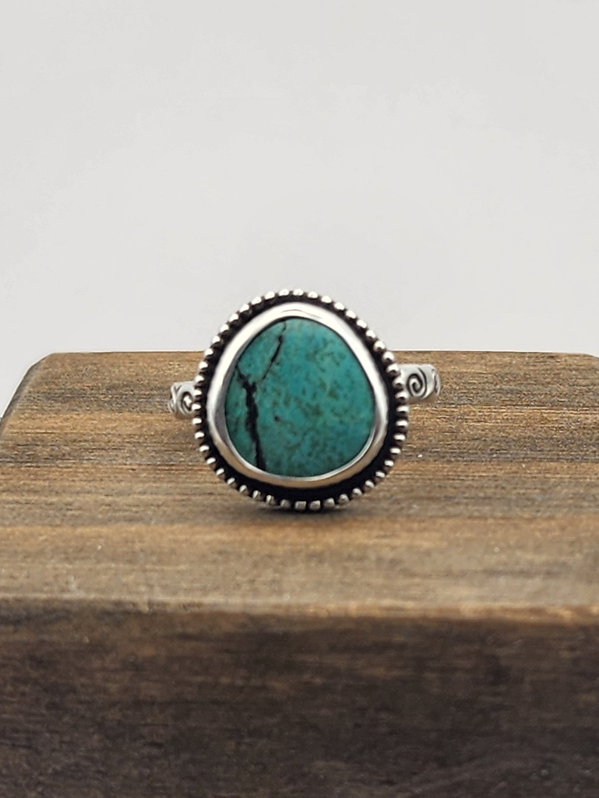 A silver ring with a round/oval turquoise stone with a beaded border displayed on a dark wooden block against a light gray background.