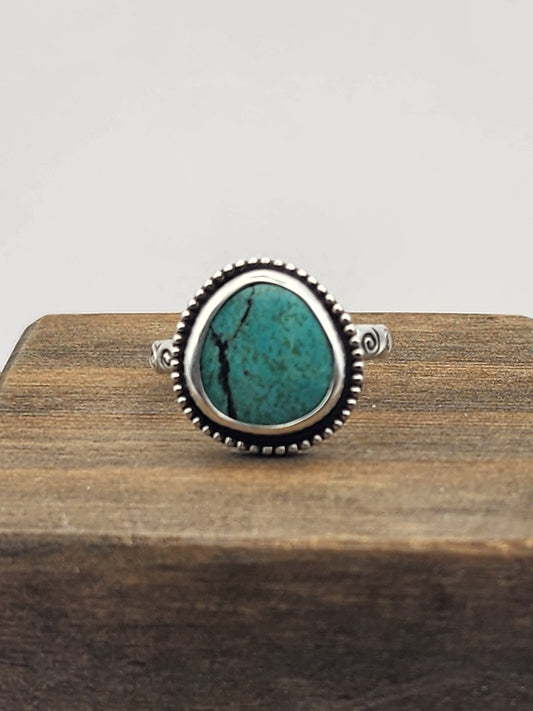 A silver ring with a round/oval turquoise stone with a beaded border displayed on a dark wooden block against a light gray background.