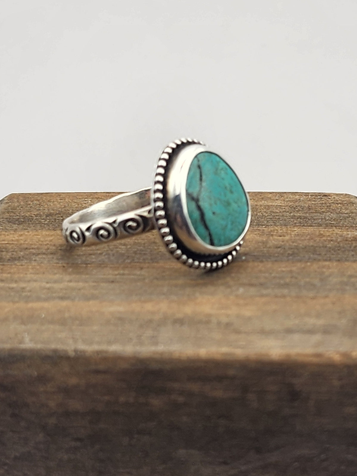 A side view of a sterling silver ring with a round/oval turquoise stone with a beaded border and swirl stamped band, displayed on a dark wooden block against a light gray background.