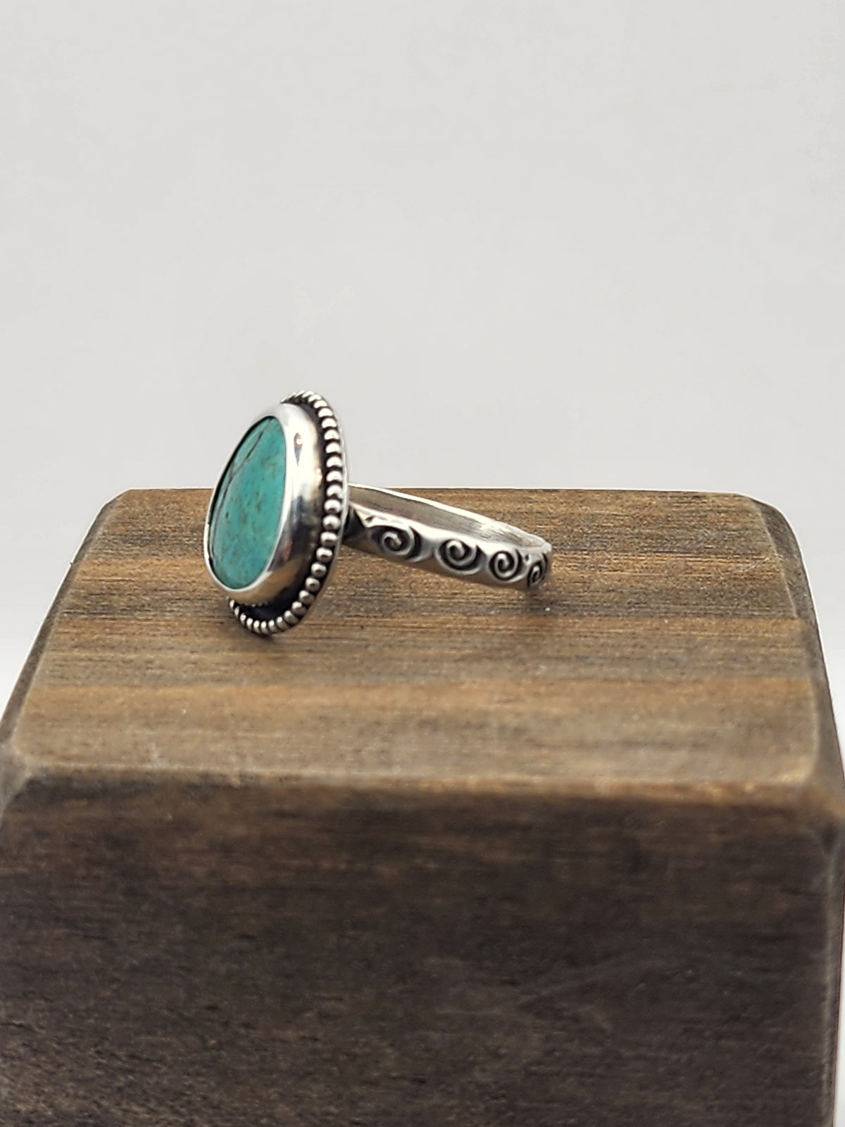 A side view of a sterling silver ring with a round/oval turquoise stone with a beaded border and swirl stamped band, displayed on a dark wooden block against a light gray background.