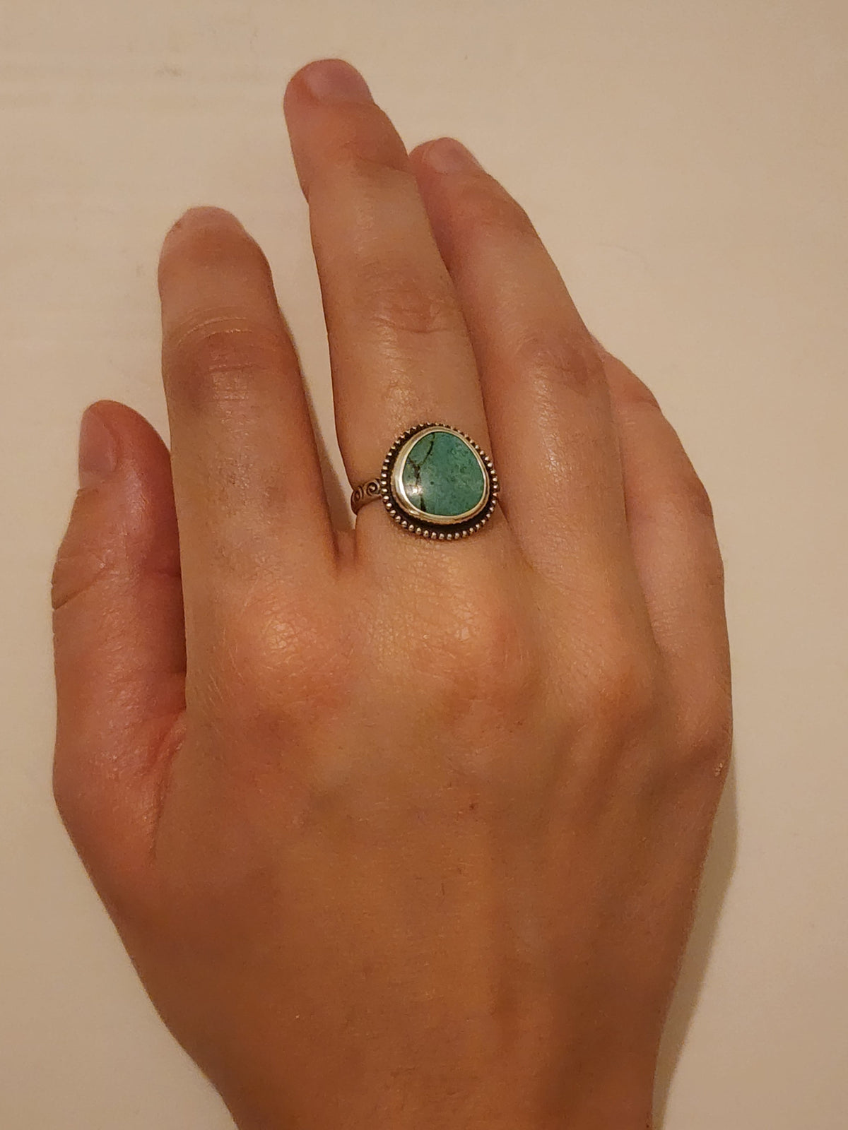 A hand wearing a sterling sliver ring with a round/oval turquoise stone, shown against a plain off-white background.