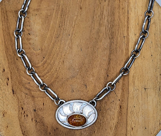 A sterling silver link chain necklace with an oval pendant featuring sunrays and an oval baltic amber in the lower center, displayed on a round wooden stand.