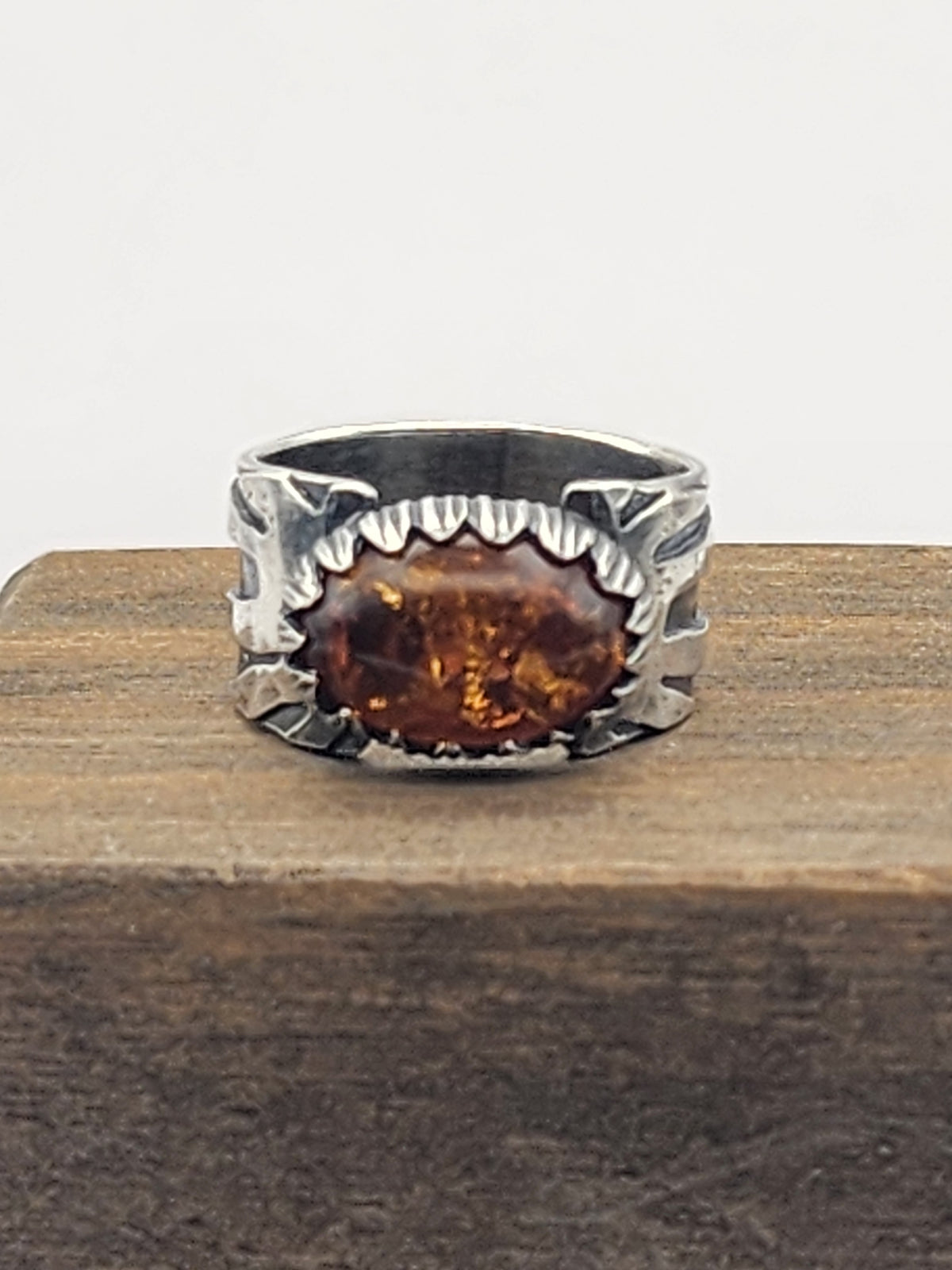 A sterling silver ring with a carved bezel and wide band and a large oval baltic amber stone in the center, displayed on a rustic wooden block against a light background.