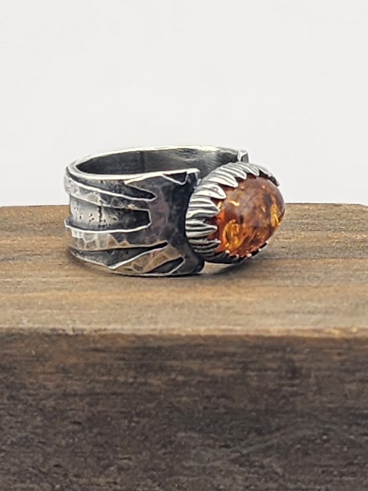 A side view of a sterling silver ring with a textured, hammered band and a large, oval baltic amber stone set in a carved silver bezel, resting on a dark wooden surface against a light gray background.