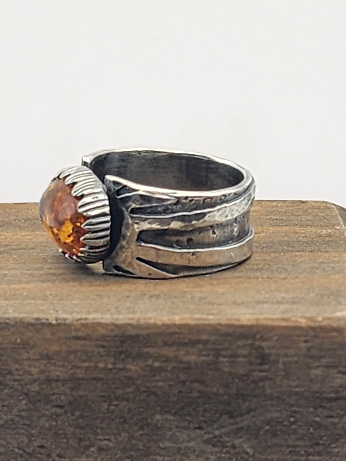 A side view of a sterling silver ring with a textured, hammered band and a large, oval baltic amber stone set in a carved silver bezel, resting on a dark wooden surface against a light gray background.