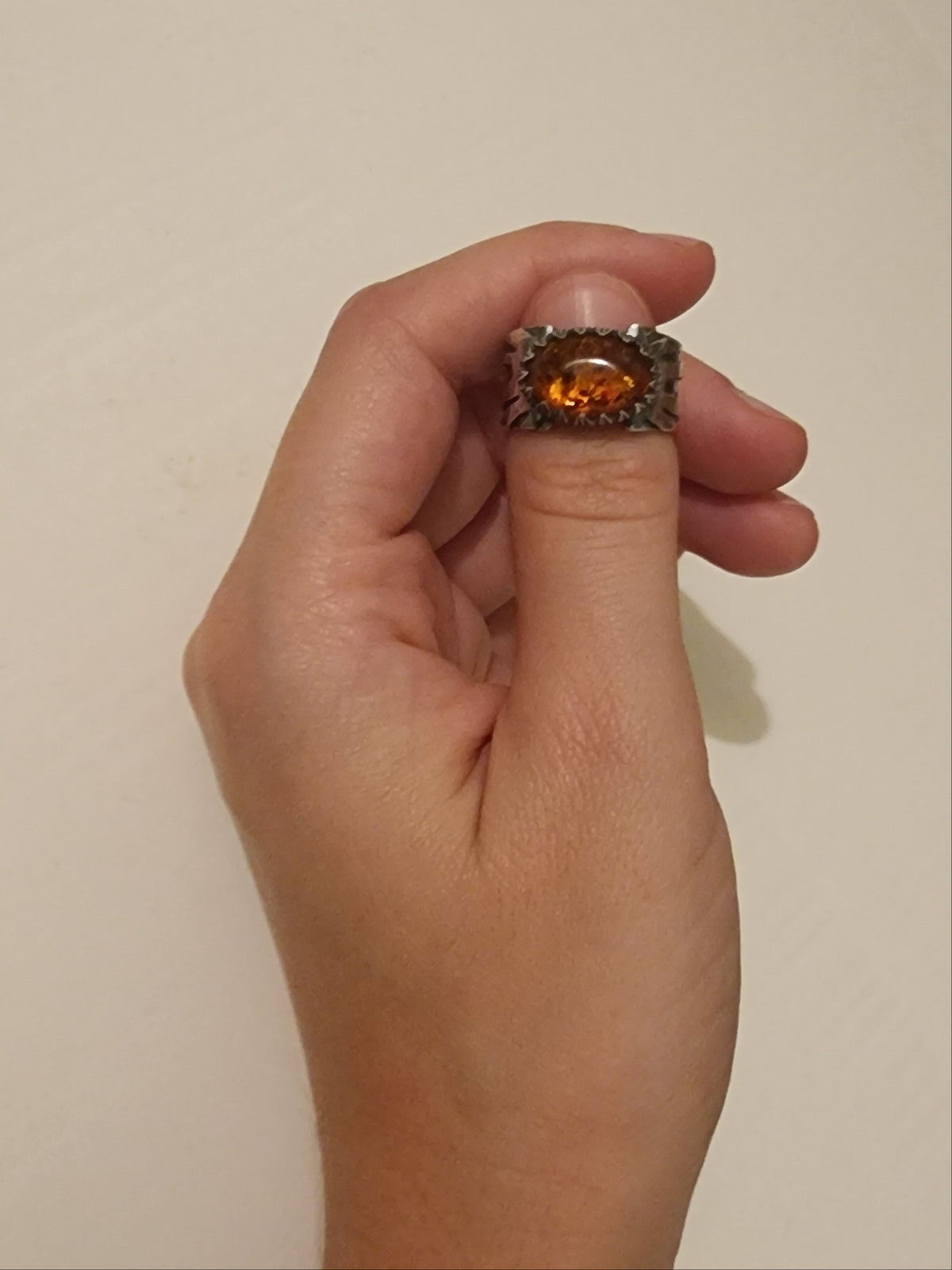 A hand holding a ring over the thumbnail. Ring is sterling silver with oval baltic amber and carved bezel on large sterling silver band, shown against a plain off-white background.