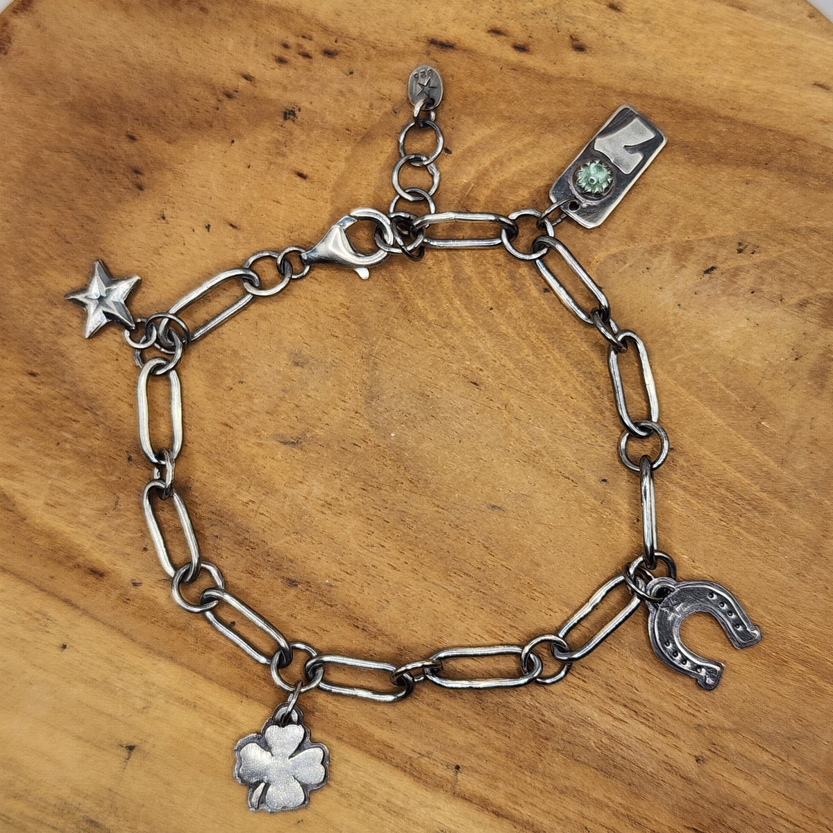 A sterling silver chain bracelet with charms, including a four-leaf clover, horseshoe, star, and a number 7 with a green gemstone, displayed on a wooden surface.