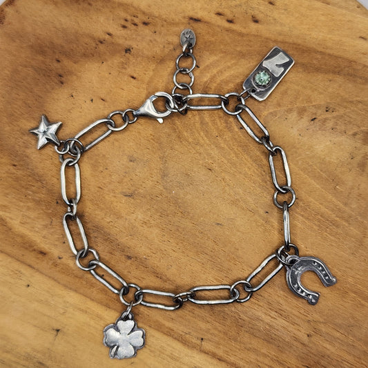 A sterling silver chain bracelet with charms, including a four-leaf clover, horseshoe, star, and a number 7 with a green gemstone, displayed on a wooden surface.