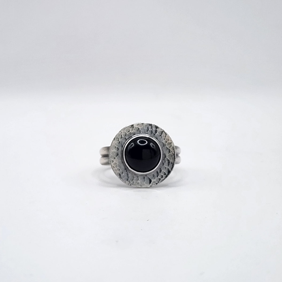 A sterling silver ring with a double band and a hammered halo surface with large round black cabochon in the center, on a white background.