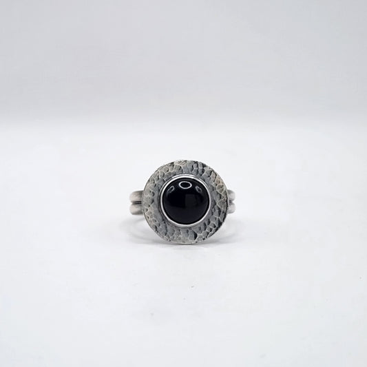 A sterling silver ring with a double band and a hammered halo surface with large round black cabochon in the center, on a white background.
