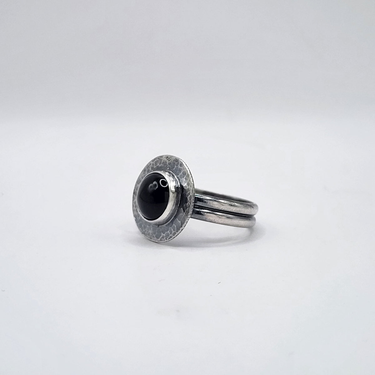 A sterling silver ring with a double band and a textured circular halo holding a black round stone, set against a white background.