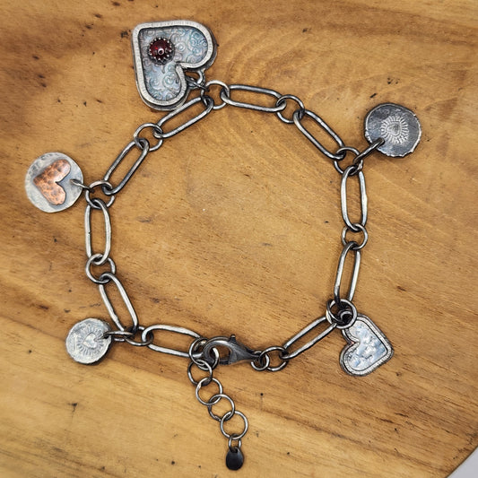 A sterling silver paperclip chain bracelet with multiple charms, including two heart charms, two round silver disc with stamped heart bursts, displayed on a round wooden board.