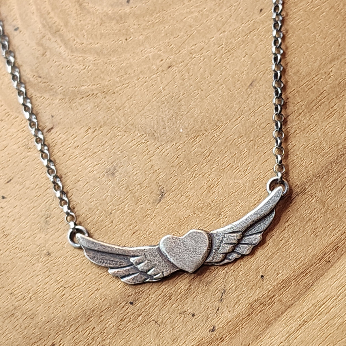 A sterling silver winged heart pendant necklace on a matching chain, displayed on a light brown wooden surface.