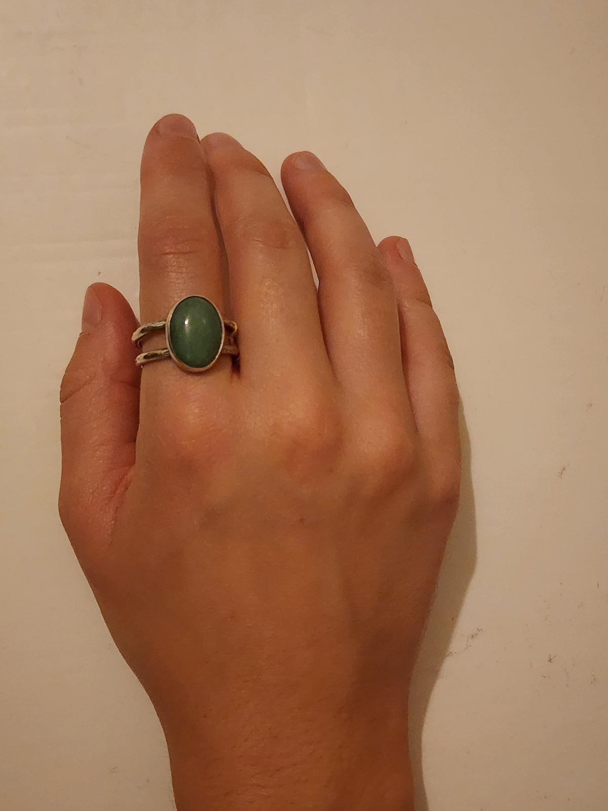A sterling silver ring with large oval green stone on the pointer finger of a hand photographed on an off white background.